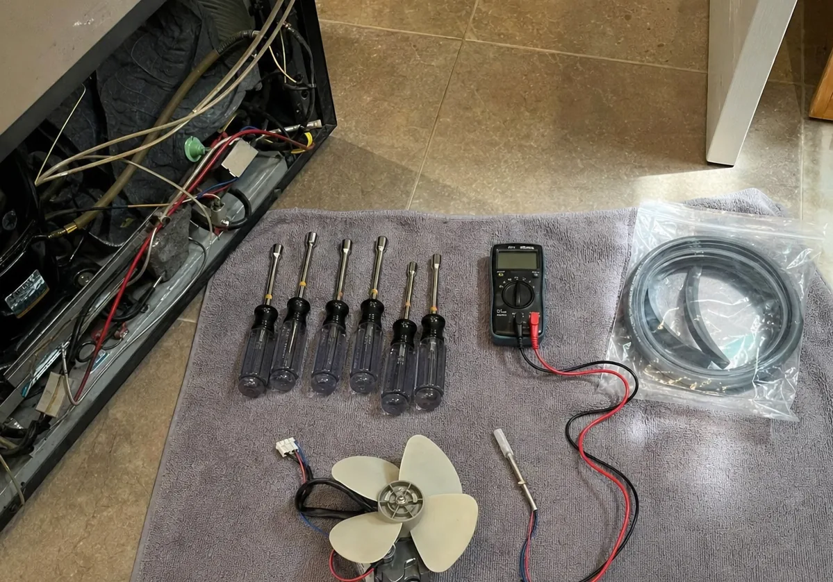 A close overhead view of nut drivers and a digital multimeter on a folded towel, with a replacement fan motor and its wiring harness set out below them.