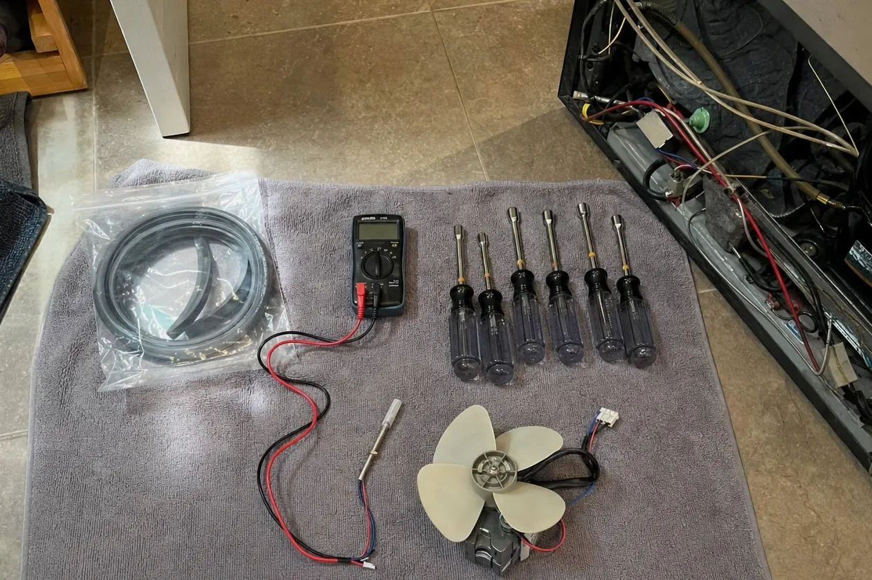 A thermistor temperature probe and its two-wire lead lying on a towel beside a replacement fan motor, with a wooden wine rack holding bottles at the edge of the frame.