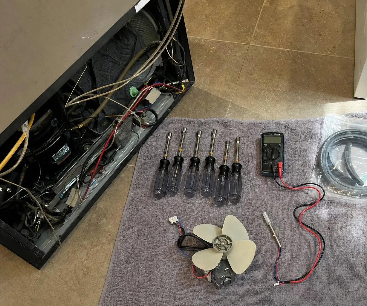 A built-in refrigerator with its lower machine compartment swung open on the left, compressor, copper lines and wiring loom in plain view, and on the floor beside it a towel laid out with nut drivers, a meter, a replacement fan motor and a bagged door gasket.