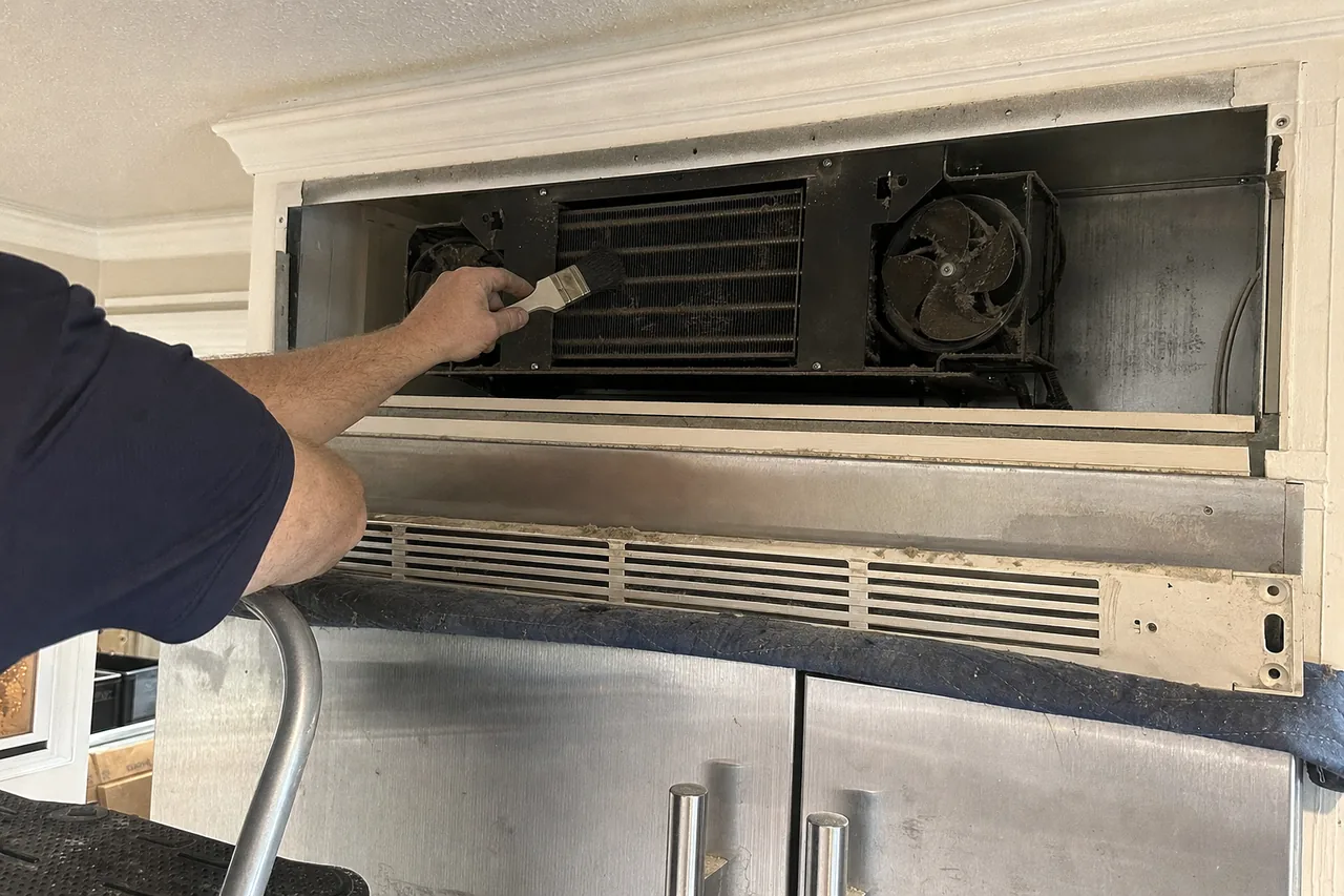 A technician reaching in with a soft brush across the fins of a condenser coil mounted above a stainless refrigerator, the removed grille panel resting to one side.