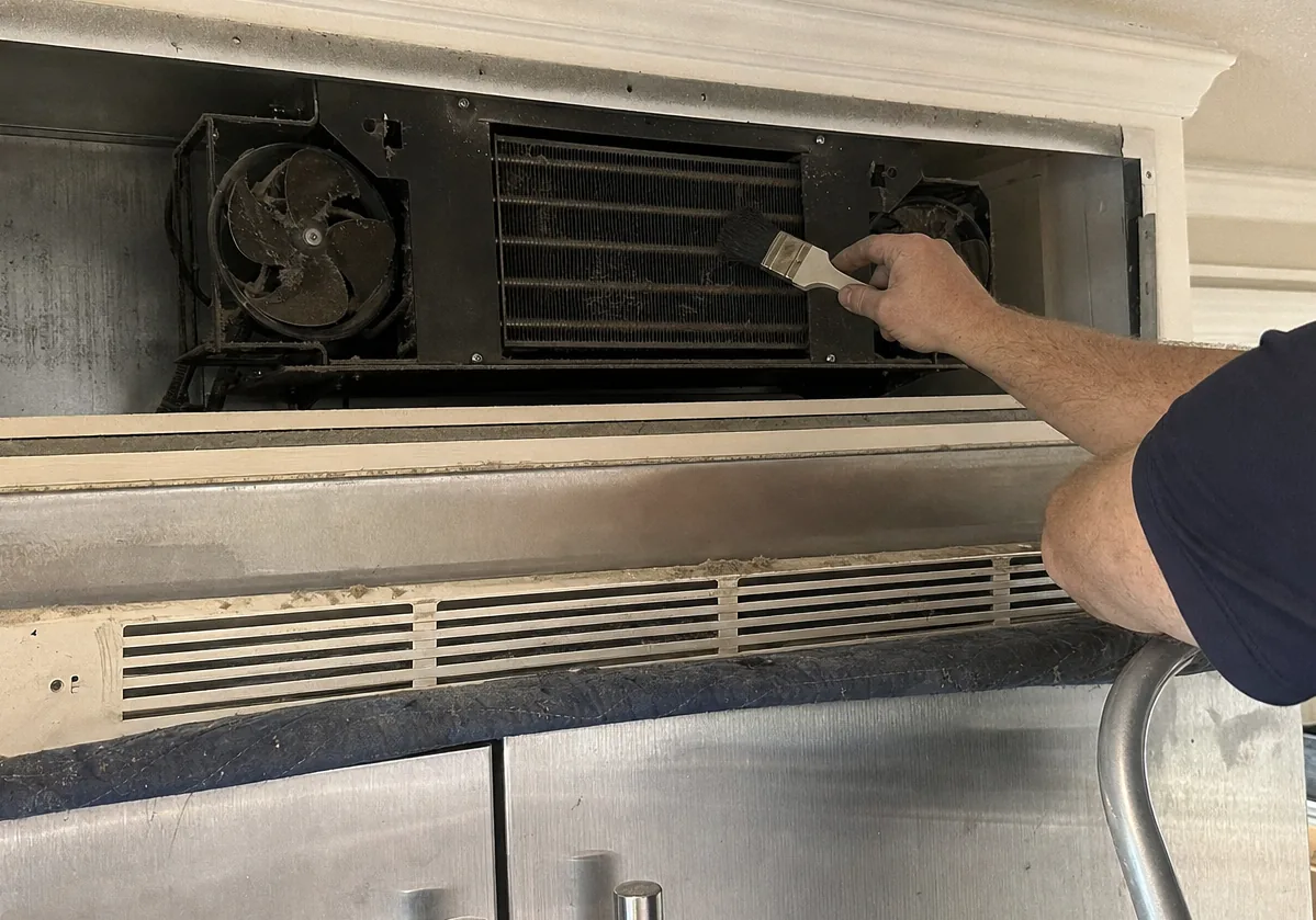 A close view of a condenser coil matted grey with lint behind the open upper grille of a built-in refrigerator, one dust-caked fan blade beside it.