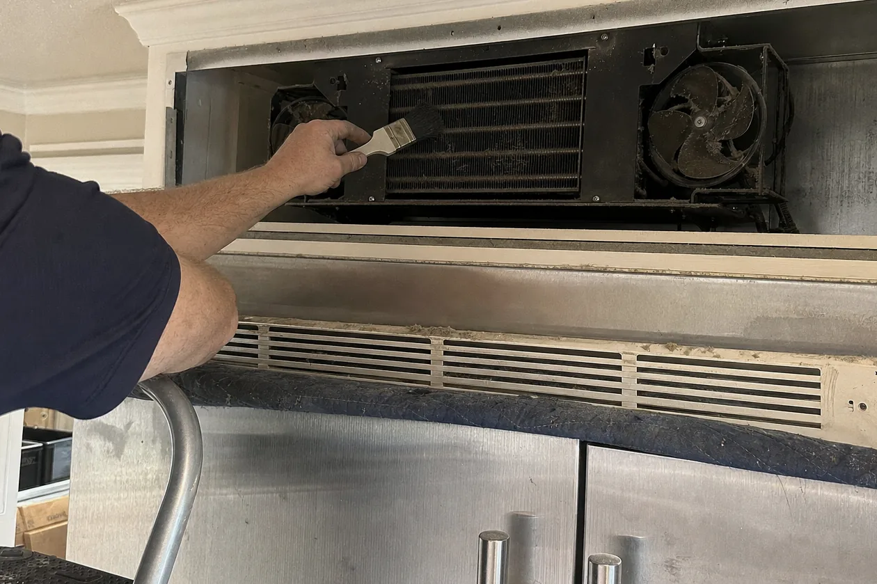 The condenser deck of a built-in refrigerator with its upper grille lifted out, twin fans and a grey-matted coil exposed, photographed on the job from a step stool.