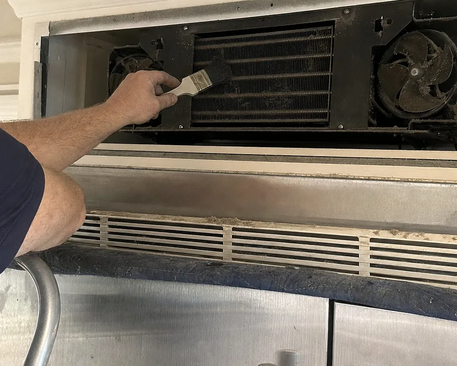 A tight view of a dust-clogged condenser coil with a soft brush drawn across the fins, the fan shroud dark with grime beside it.